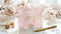 Pink sorry card with gold calligraphy text surrounded by cherry blossoms and a golden pen on a white table, apology concept.