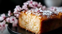 Homemade vanilla pound cake slice dusted with powdered sugar, decorated with delicate pink cherry blossom branches against dark background.