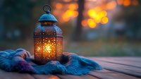 Glowing oriental lantern with ornate metalwork on wooden surface against blurred sunset bokeh, cozy atmosphere created by warm light and blue textile fabric.