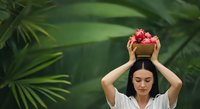 Young woman with dark hair and fair complexion holding a golden bowl filled with pink flowers above her head against lush green tropical foliage background. Suitable for wellness, beauty, and lifestyle marketing.