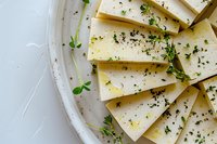 Fresh cheese slices with herbs and black pepper on white plate, gourmet dairy product for culinary presentations and food photography.