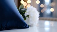 Elegant navy blue silk pillow with white peony flower against blurred bokeh lights background, creating romantic and luxurious home decor atmosphere.