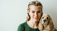 Woman with blonde braided hair wearing green shirt posing with golden cocker spaniel dog against white background for pet adoption or veterinary marketing.