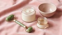 Natural skincare routine with jade roller, cream jar, and ceramic bowl on soft pink fabric for beauty and wellness rituals.