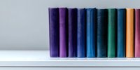 Row of colorful hardcover books standing on white shelf against light wall, spines showing gradient from purple through blue to orange, minimal library setup.