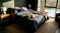 Luxurious bedroom interior with unmade navy silk bedding, gold accents, wooden nightstands, and large windows overlooking nature for home decor and lifestyle design projects.