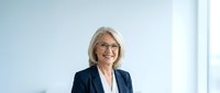Mature woman with gray hair and glasses wearing dark blazer and white shirt, smiling at camera in bright office setting for corporate portraits and business headshots.