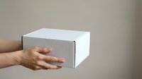 Male hand holding white cardboard box on neutral background, suitable for product packaging mockups, shipping logistics, delivery services, and e-commerce presentations.