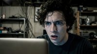 Young man with curly dark hair and fair complexion working late at laptop in cluttered workspace, appearing stressed or focused on screen for tech, programming, or business projects.