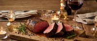 Sliced roast beef tenderloin on wooden cutting board with wine glass and candles creating elegant dining atmosphere for holiday celebrations.