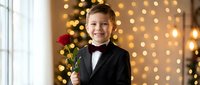 Young boy in formal black tuxedo holding red rose with warm Christmas tree lights bokeh background creating festive holiday atmosphere for celebrations.