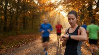 Young athletic woman checking fitness tracker during autumn morning jog in park with golden sunlight filtering through colorful fall foliage and runners.