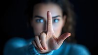 Young woman with intense blue eyes gesturing with index finger in dark atmospheric setting, focus on hand in foreground while face remains mysteriously blurred.