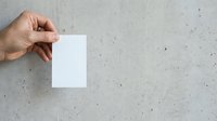 Adult hand with medium skin tone holding blank white card against textured concrete wall background for branding, mockup, and design presentation purposes.