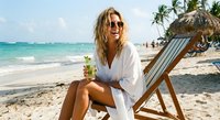 Young blonde woman in white beach cover-up relaxing on striped deck chair with tropical drink at paradise resort with palm trees and turquoise ocean water.