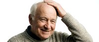 Senior man with white hair and light complexion, wearing gray turtleneck sweater, smiling warmly while touching head with hand in relaxed pose for portrait and lifestyle content.