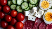 Fresh ingredients for healthy salad with boiled eggs, cherry tomatoes, sliced cucumbers, feta cheese cubes and raw meat arranged in rows on dark background.