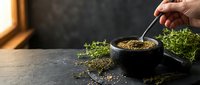 Hand sprinkling herbs and spices into dark bowl on slate surface with fresh greenery for culinary seasoning and cooking preparation.