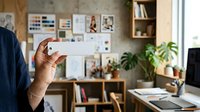 Young adult male with medium brown skin tone holding blank white card in modern home office workspace with wooden shelving, plants, and design mood board on wall.