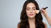 Professional makeup artist applying foundation with brush to young brunette woman with closed eyes and wavy hair against neutral background for beauty salon.