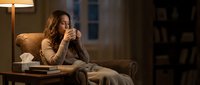 Woman with long brown hair, Caucasian ethnicity, sitting on brown sofa in warm-lit home office, resting chin on hand in thoughtful pose, wearing beige cardigan, contemplative mood.