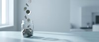 Coins falling into glass jar on white table in bright modern kitchen interior for savings and financial planning concepts.
