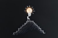 Chalk drawing on blackboard showing stick figure with glowing lightbulb head standing on top of stairs, representing achievement and bright ideas in business.