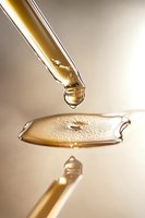 Golden liquid drop falling from glass dropper onto reflective surface with water ripple effect, captured in macro photography with soft beige background.