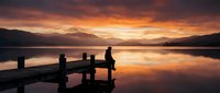 Silhouette of person sitting on wooden dock at sunrise over calm lake with mountains, reflecting golden light on water surface for nature and travel concepts.