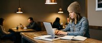 Young woman with beanie hat working on laptop in modern coffee shop cafe. Remote work, freelancing, digital nomad lifestyle concept for business content.