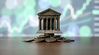 Miniature bank building with classical columns standing on pile of coins with blurred financial charts background representing banking investment growth.