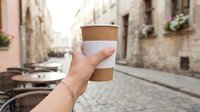 Hand holding takeaway coffee cup with kraft sleeve on European street with historic architecture and bicycle in background.