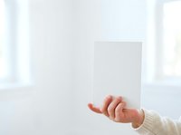 Hand holding blank white card or paper against bright white background with natural light, suitable for mockup, branding, greeting card design, and promotional material presentations.