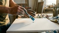 Artist hands using blue acrylic paint tube on white canvas in creative studio workspace with brushes and art supplies for painting project.