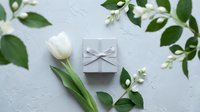 Elegant white tulip and gray gift box with ribbon surrounded by green leaves on light textured background, flat lay for spring holiday.