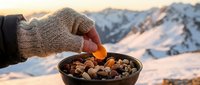 Young adult wearing knit gloves holding dried fruit over bowl of mixed nuts with snowy mountain landscape in background. Winter hiking snack.