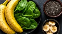 Fresh ripe bananas, green spinach leaves, and chia seeds in bowls on dark background. Healthy superfood ingredients for smoothie preparation.
