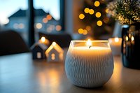 Glowing aromatic candle in white textured ceramic holder creates cozy atmosphere with decorative houses and bokeh lights in background for holiday decor.