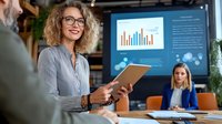 Caucasian businesswoman with curly hair and glasses holding tablet during corporate presentation, with data charts on screen and colleague in background.
