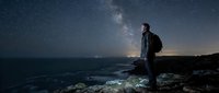 Man standing on rocky coastal cliff at night gazing at starry sky and Milky Way galaxy over ocean. Nighttime astronomy and stargazing concept.