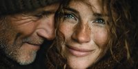 Loving mature couple sharing intimate moment together, man with beard embracing smiling woman with freckles and curly hair in warm natural lighting.