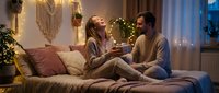 Young couple enjoying intimate conversation on cozy bed in warmly lit bedroom with fairy lights and bohemian decor creating romantic atmosphere at home.