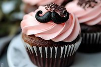 Chocolate cupcake with pink buttercream frosting topped with black mustache decoration and chocolate sprinkles, served in striped paper liner on blurred background.