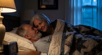 Senior Caucasian couple sharing intimate moment in bed at night, elderly man and woman embracing under cozy blanket in dimly lit bedroom for romance concepts.