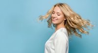 Happy blonde woman with flowing hair in white shirt against blue background expressing joy and freedom with closed eyes and genuine smile.