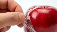 A hand removing plastic wrap from a fresh red apple against a white background, showing food packaging and produce handling.