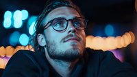 Young Caucasian man wearing glasses gazes upward thoughtfully against blurred city lights at night, creating atmospheric mood with orange and blue bokeh background.
