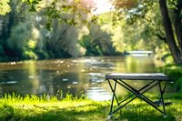 Portable camping chair on green grass by tranquil river with sunlit water and forest background, creating peaceful outdoor recreation scene.
