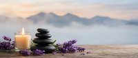 Zen spa composition with stacked black stones, burning candle and lavender flowers on wooden surface against misty mountain landscape at sunset for relaxation.