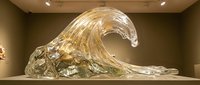 Large transparent glass wave sculpture displayed in modern art gallery with warm lighting and neutral walls creating elegant contemporary exhibition space.